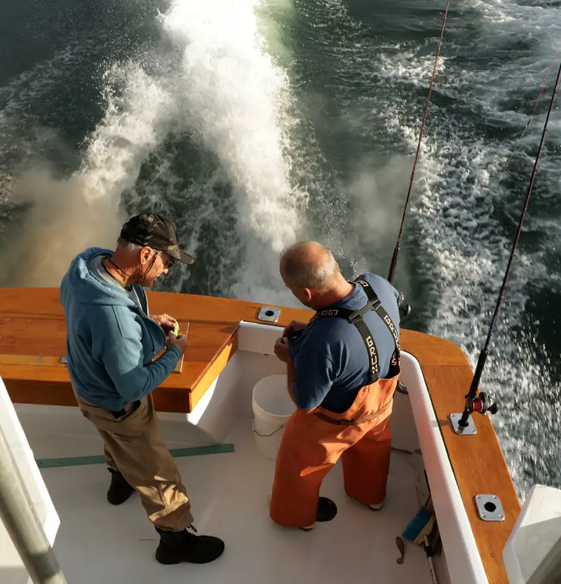 Two fishermen on a boat, preparing gear with a scenic view of waves splashing behind them under warm sunlight.