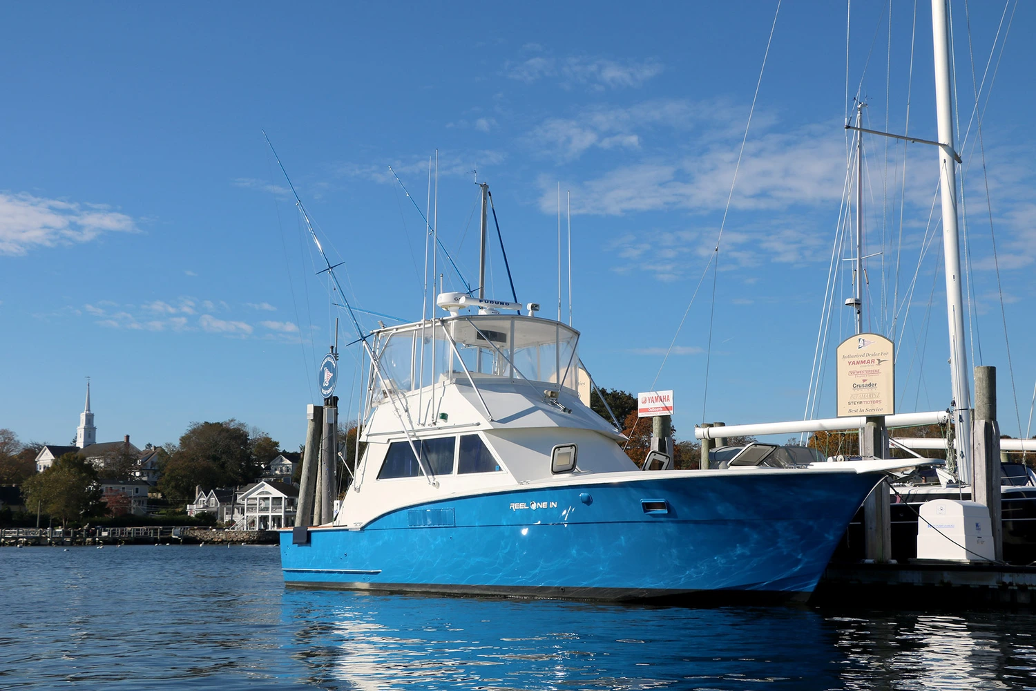 A blue fishing boat named "Reel One In" docked at a marina under a clear blue sky, with buildings and a church steeple in the background.