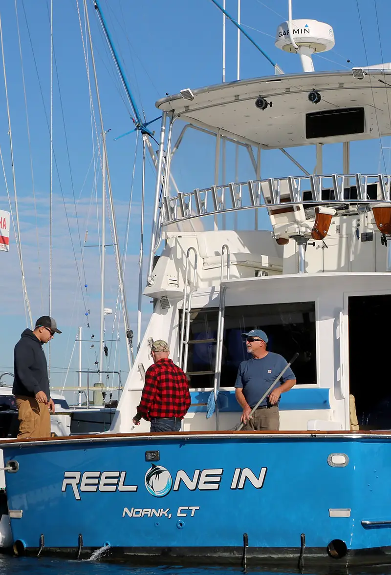 Three people are on a fishing boat named "Reel One In," docked in a marina with clear blue skies and rigging in the background.
