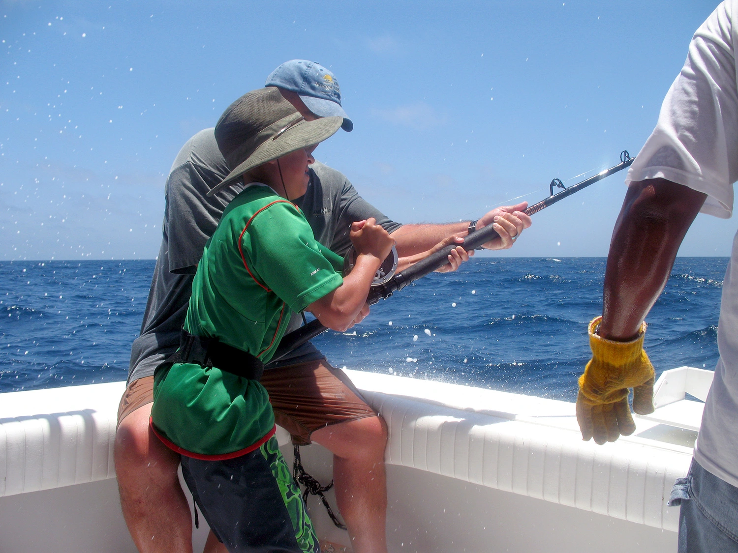 A child in a green shirt holding a fishing rod, assisted by an adult, on a boat surrounded by splashing water and blue ocean