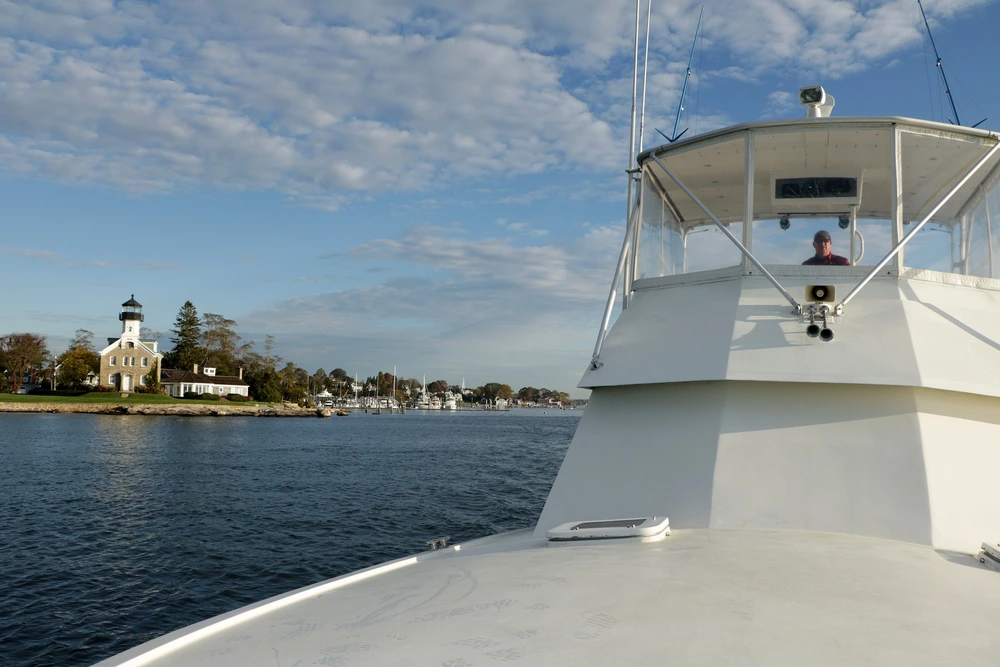 A captain steers a boat from the flybridge, surrounded by a scenic waterfront and cloudy sky. A captain steers a boat from the flybridge, surrounded by a scenic waterfront and cloudy sky.