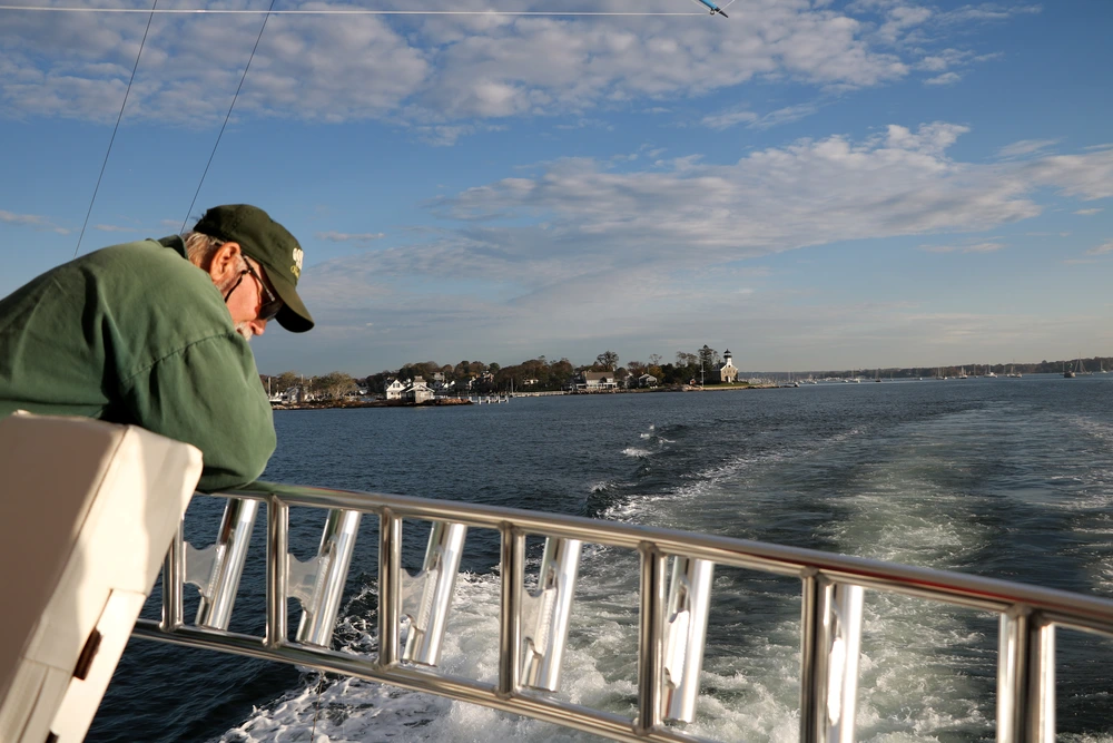 A man in a green jacket and cap leans over the side of a boat, gazing at the water and distant shoreline under a blue sky. A man in a green jacket and cap leans over the side of a boat, gazing at the water and distant shoreline under a blue sky.