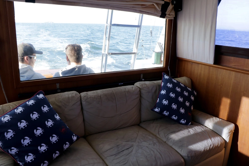 A cozy boat interior features a beige couch and crab-patterned pillows, with a view of ocean waves through a large window. A cozy boat interior features a beige couch and crab-patterned pillows, with a view of ocean waves through a large window.